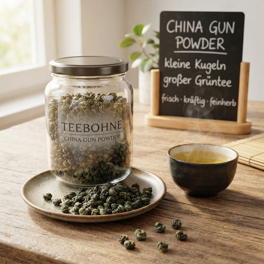 Produktfoto von TeeBohne Einbeck: Ein Glasgefäß gefüllt mit China Gun Powder Grünteekügelchen, daneben eine kleine Schale mit frisch aufgebrühtem Grüntee und eine handbeschriebene Kreidetafel auf einem Holztisch, in einer hochwertigen und authentischen Teefachhandels-Atmosphäre.
