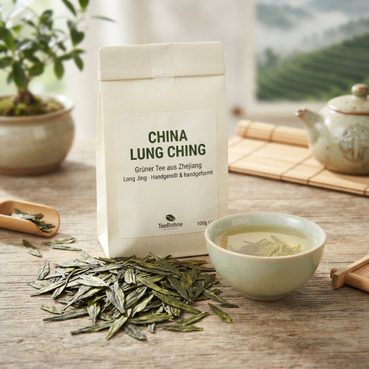 TeeBohne: Eine elegante Teeverpackung mit der Aufschrift „CHINA LUNG CHING – Grüner Tee aus Zhejiang“, umgeben von flachen, handgerollten Teeblättern und einer Teeschale mit hellgelbem Aufguss auf einer Holzoberfläche