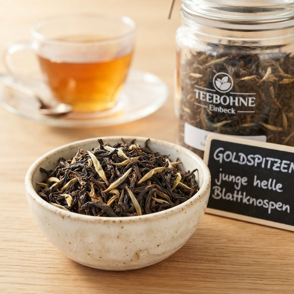 TeeBohne Einbeck: Eine kleine Keramikschale im Vordergrund zeigt dunkle Assam-Teeblätter mit gut erkennbaren, hellen goldfarbenen Blattknospen (Goldspitzen). Daneben stehen ein transparentes Teeglas mit derselben Mischung und eine kleine Kreidetafel mit der Aufschrift "GOLDSPITZEN junge helle Blattknospen", während im Hintergrund eine Tasse kräftiger Ostfriesentee unscharf zu sehen ist